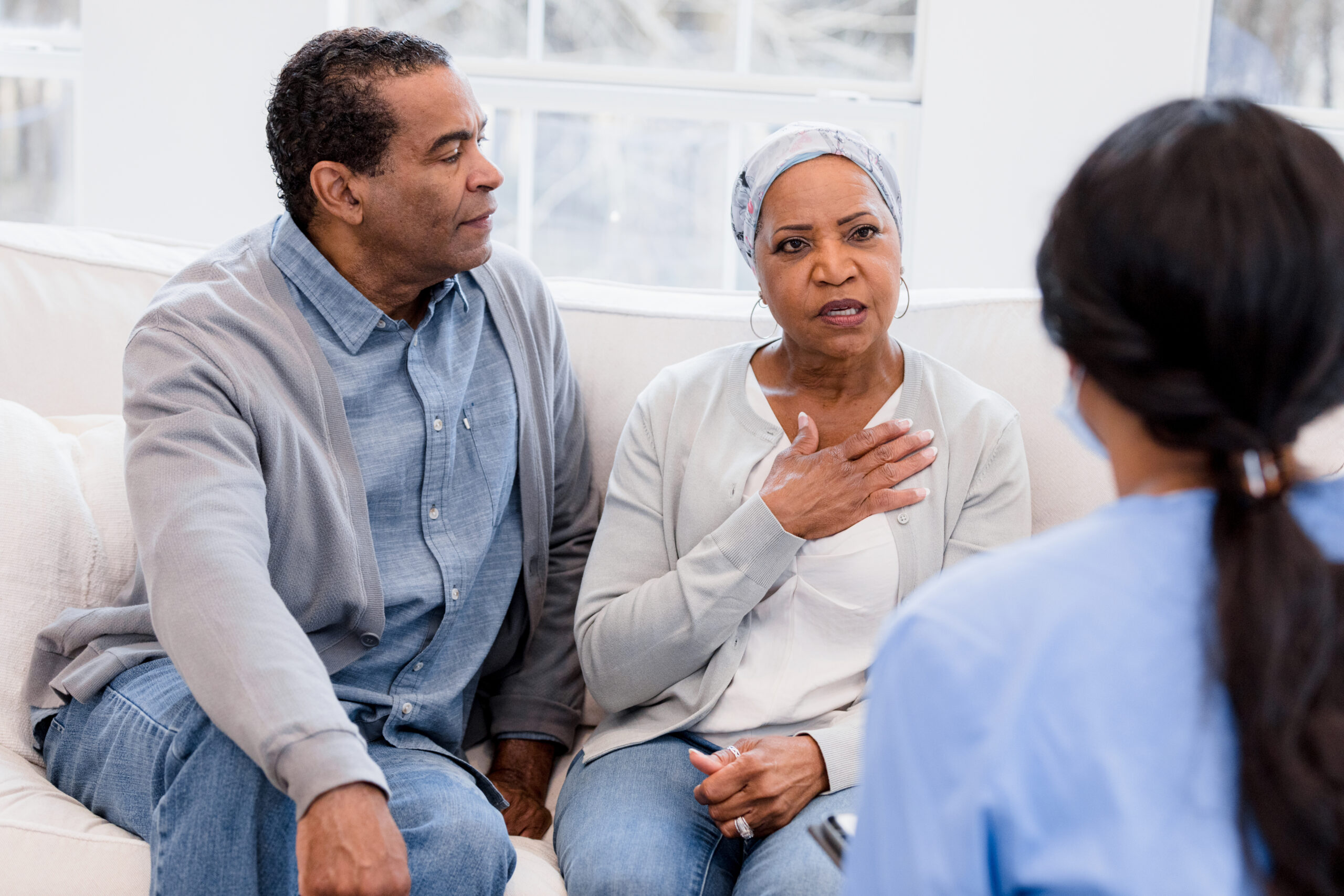 An older black couple sitting on a couch with the senior woman holds her chest and talking to a caregiver