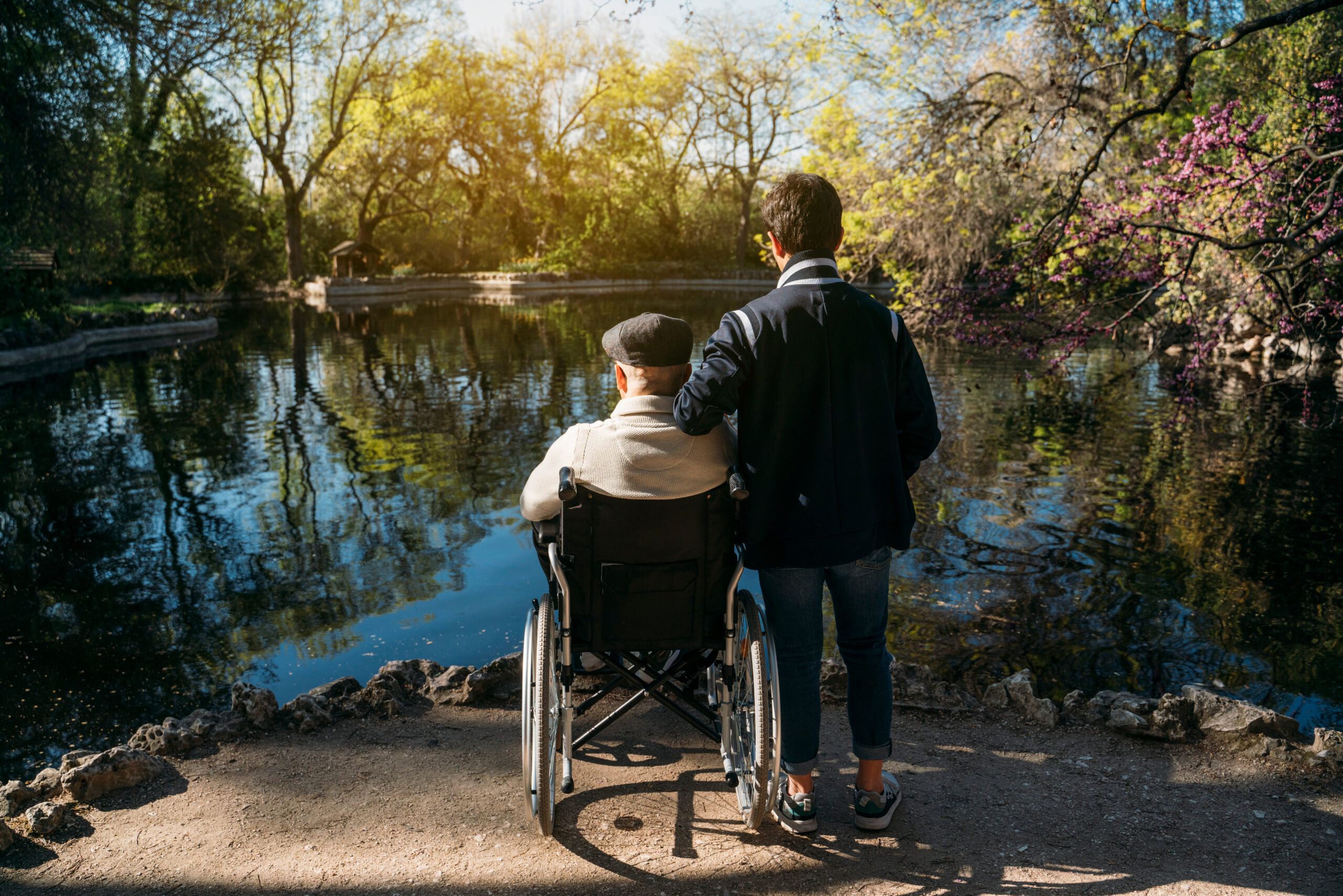 Older man in wheelchair with caregiver looking over a pond
