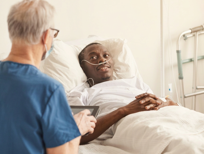 A black man laying in a hospital bed is talking with his gray haired doctor